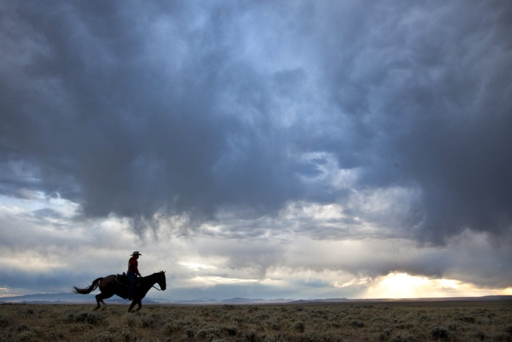 A lone cowboy rides a horse across a vast, open plain under a dramatic, stormy sky in Wind River Country.