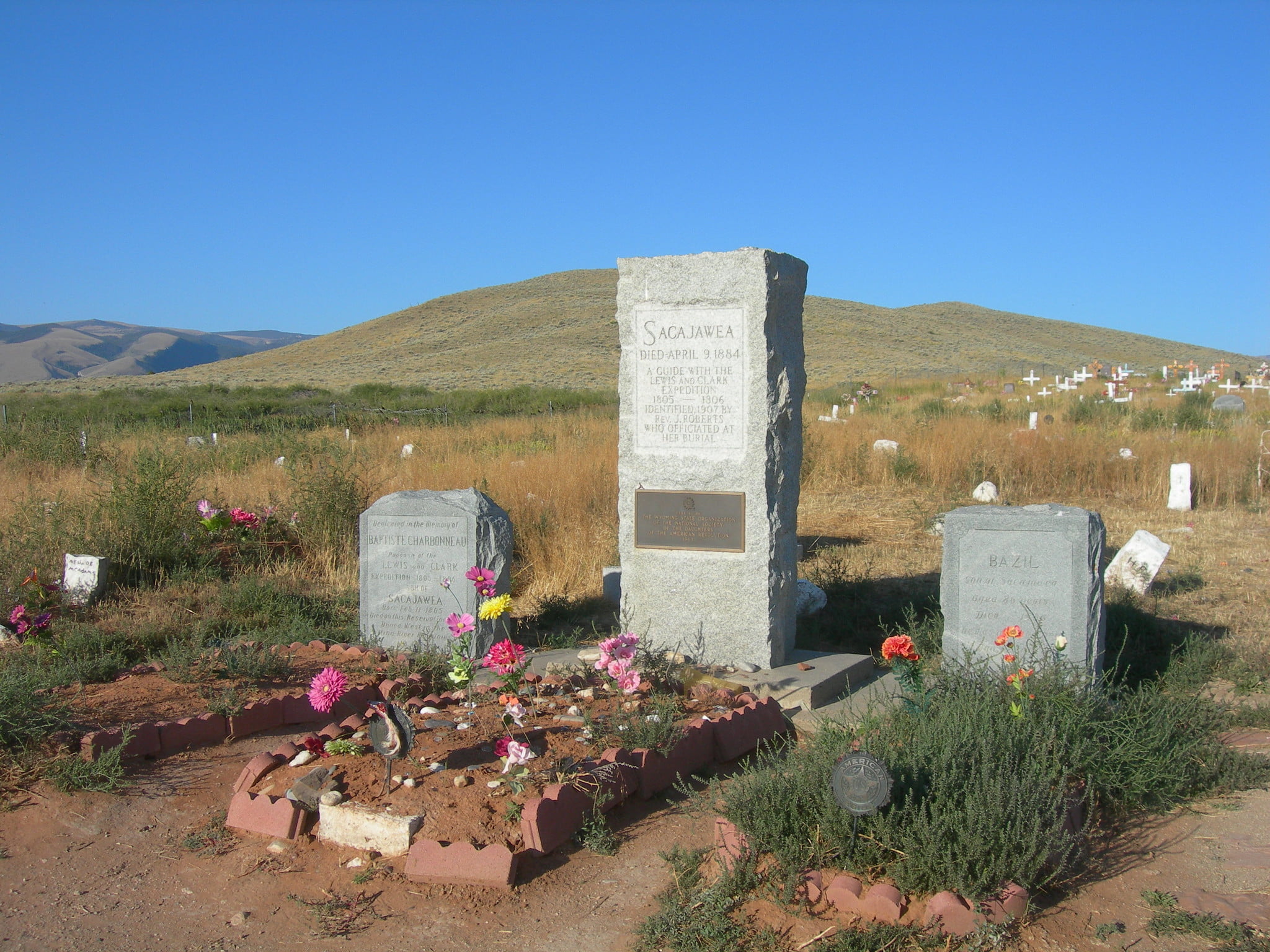 A serene cemetery with a prominent stone monument surrounded by smaller headstones, all adorned with colorful flowers against a backdrop of rolling hills and a clear blue sky in Wind River Country.