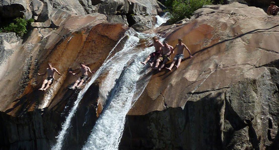 Four young men are seen sliding down a steep, rocky waterfall, enjoying the thrill of the water rushing beneath them in Wind River Country.