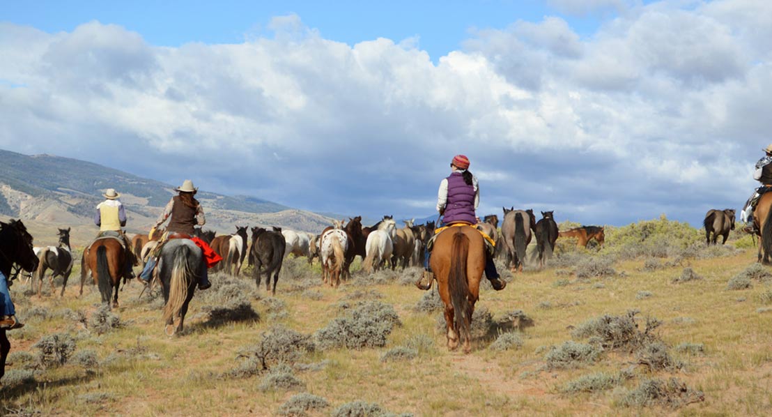 A group of riders on horseback traverse a vast, open plain dotted with wild horses under a partly cloudy sky in Wind River Country.