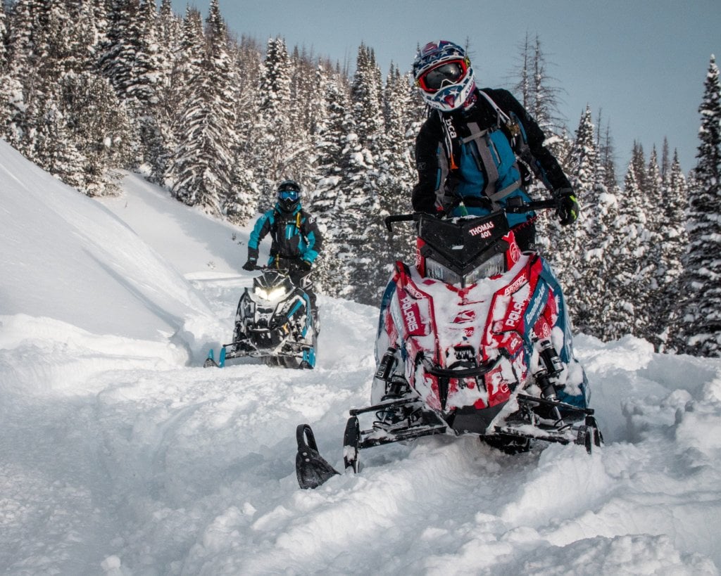 Two snowmobilers navigate through a snowy forest, one on a red and blue snowmobile and the other on a black and blue one, both wearing protective gear in Wind River Country.