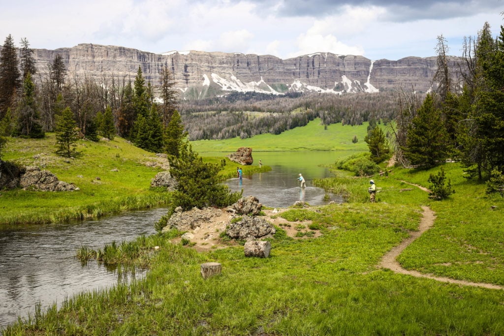 A serene mountain landscape with a winding river, lush green meadows, and towering peaks in the distance in Wind River Country.