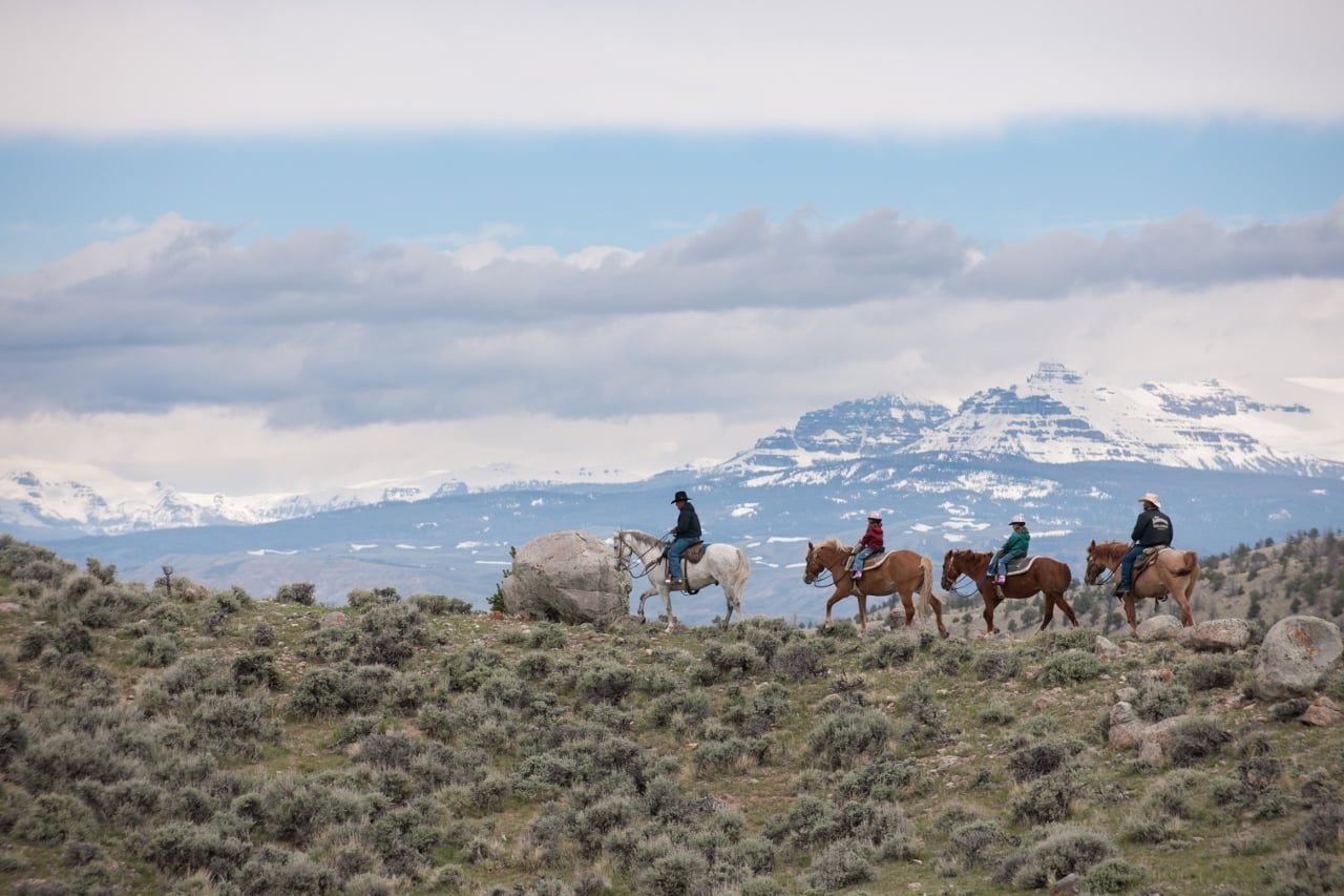 Four riders on horseback traverse a grassy hillside with a backdrop of snow-capped mountains and a partly cloudy sky in Wind River Country.