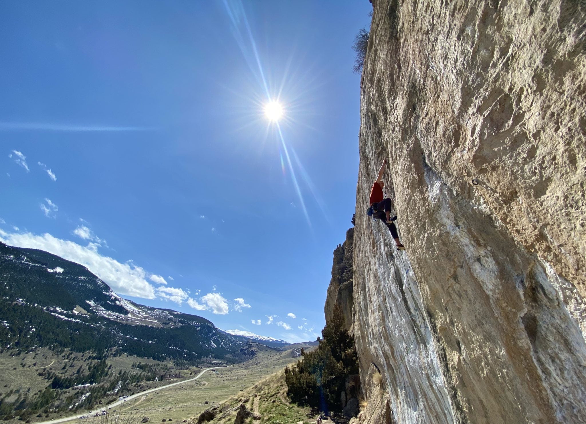A rock climber ascends a towering cliff face under a bright sun, with snow-capped mountains and a winding road visible in the distance in Wind River Country.