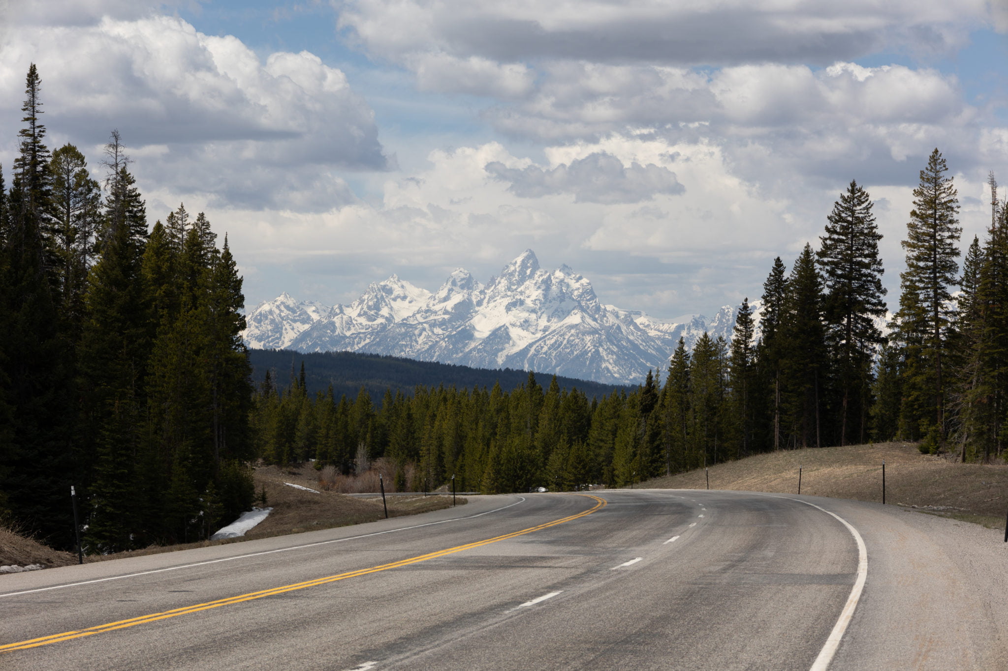 A winding road cuts through a lush green forest, leading towards majestic snow-capped mountains under a partly cloudy sky in Wind River Country.