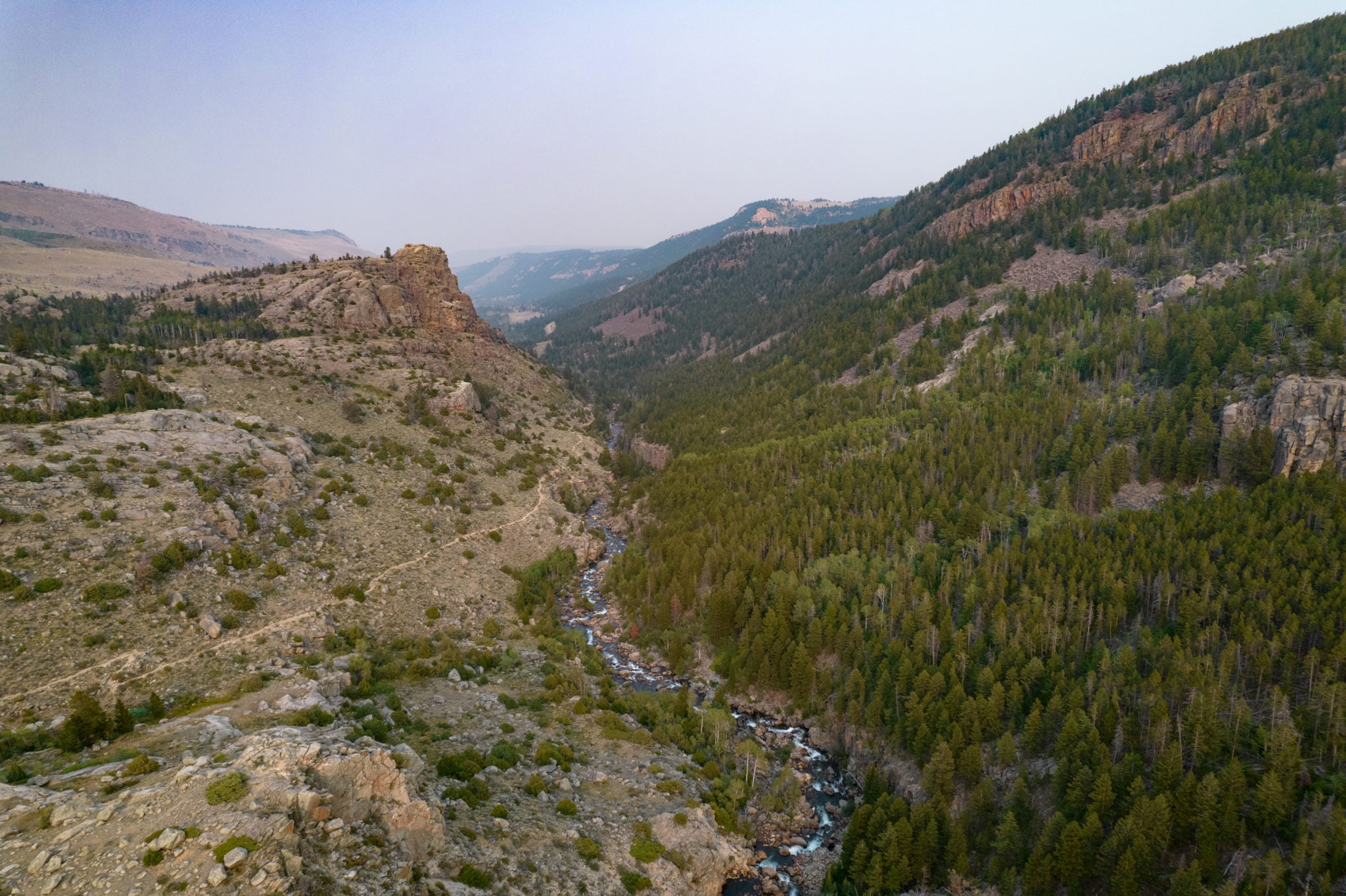 A panoramic view of a winding river cutting through a lush green valley, flanked by rugged mountains and dense forests in Wind River Country.