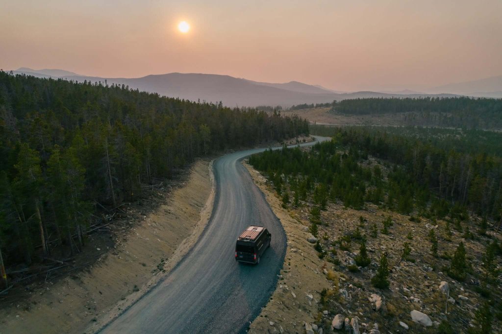 A winding dirt road cuts through a vast forest, leading towards distant mountains under a setting sun in Wind River Country.