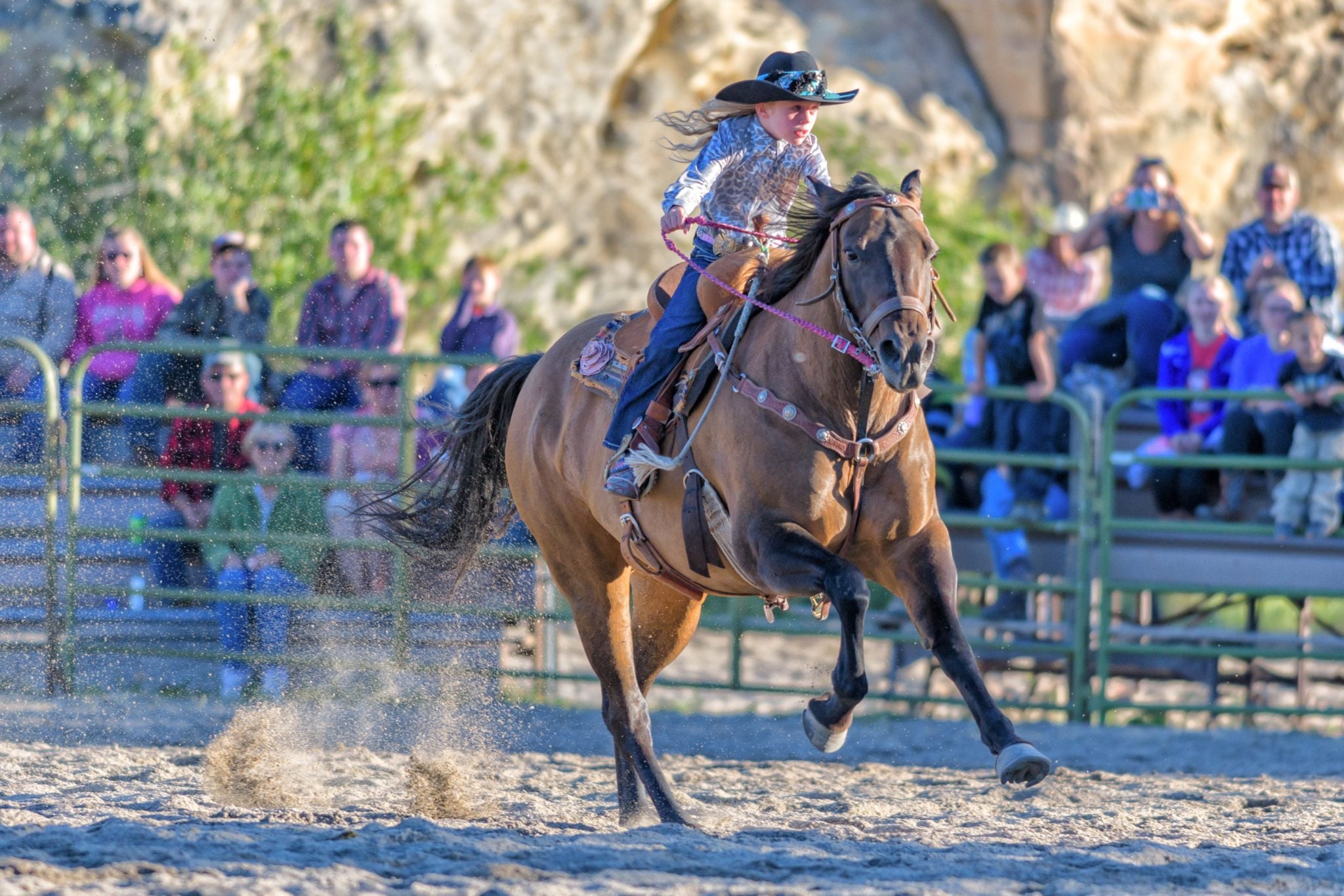 A young girl in a cowboy hat confidently rides a bucking horse in a rodeo arena, with an excited crowd watching from the stands in Wind River Country.