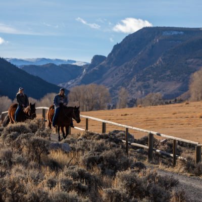Two riders on horseback enjoy a leisurely ride along a trail with a panoramic view of a valley and distant mountains in the backdrop.