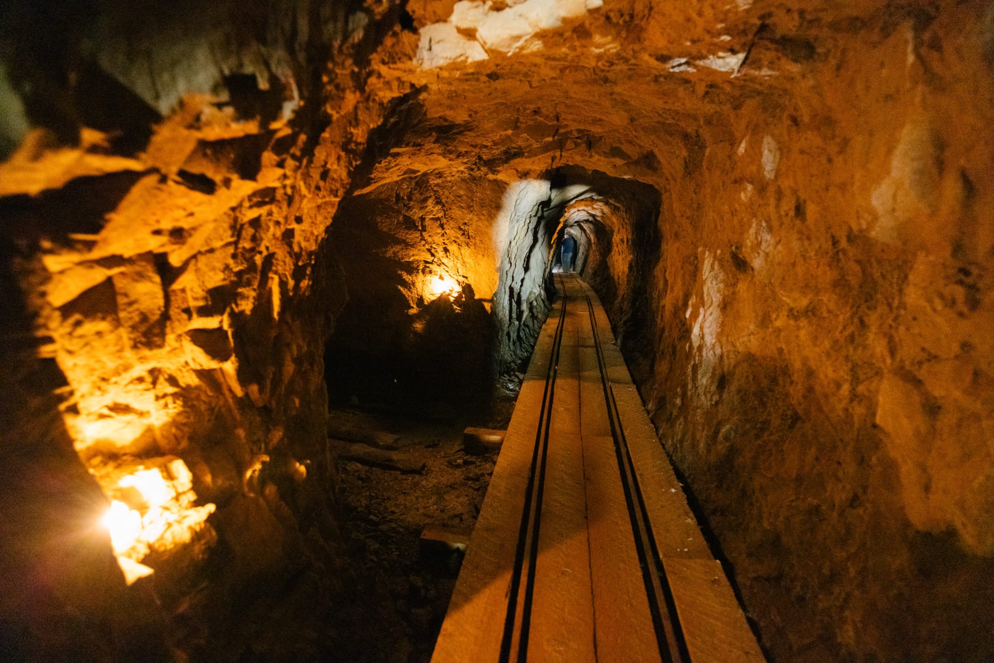 A narrow wooden track winds through a dimly lit, rocky tunnel with flickering lights along the walls in Wind River Country.