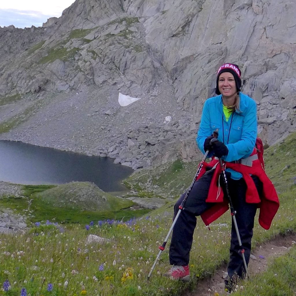 A hiker stands on a mountain trail, smiling, with a serene alpine lake and rugged peaks in the background in Wind River Country.