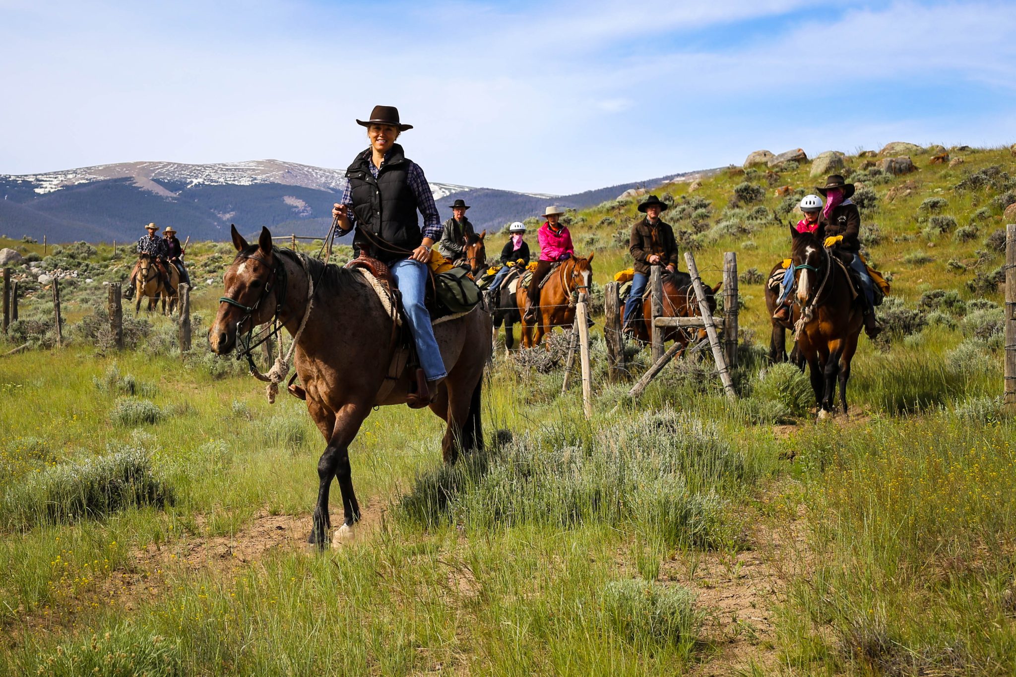 A group of riders on horseback traverse a grassy field with a backdrop of snow-capped mountains in Wind River Country.