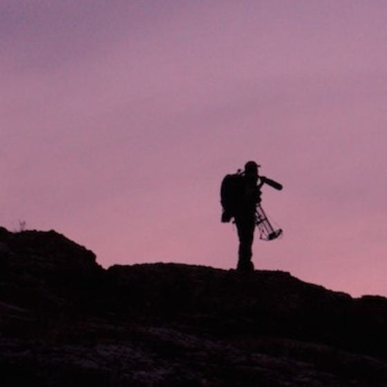 A silhouette of a person with a backpack and a tripod stands on a rocky outcrop at sunset in Wind River Country.
