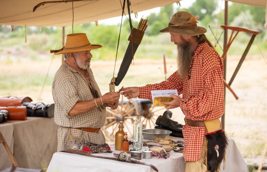 Two men dressed in period clothing engage in a transaction at a frontier market stall, surrounded by various goods and tools in Wind River Country.