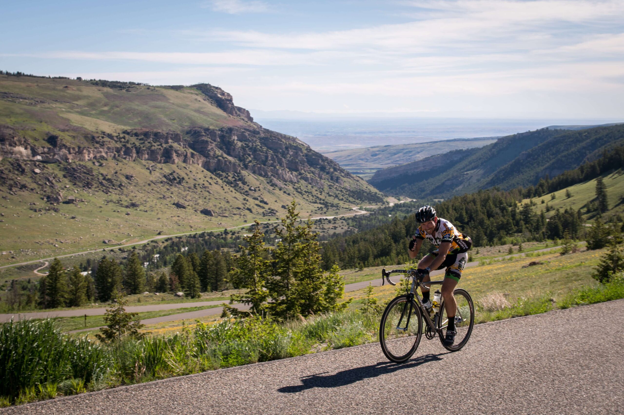 A cyclist in a yellow and black outfit vigorously pedals up a scenic mountain road, surrounded by lush greenery and dramatic rock formations, under a clear blue sky.