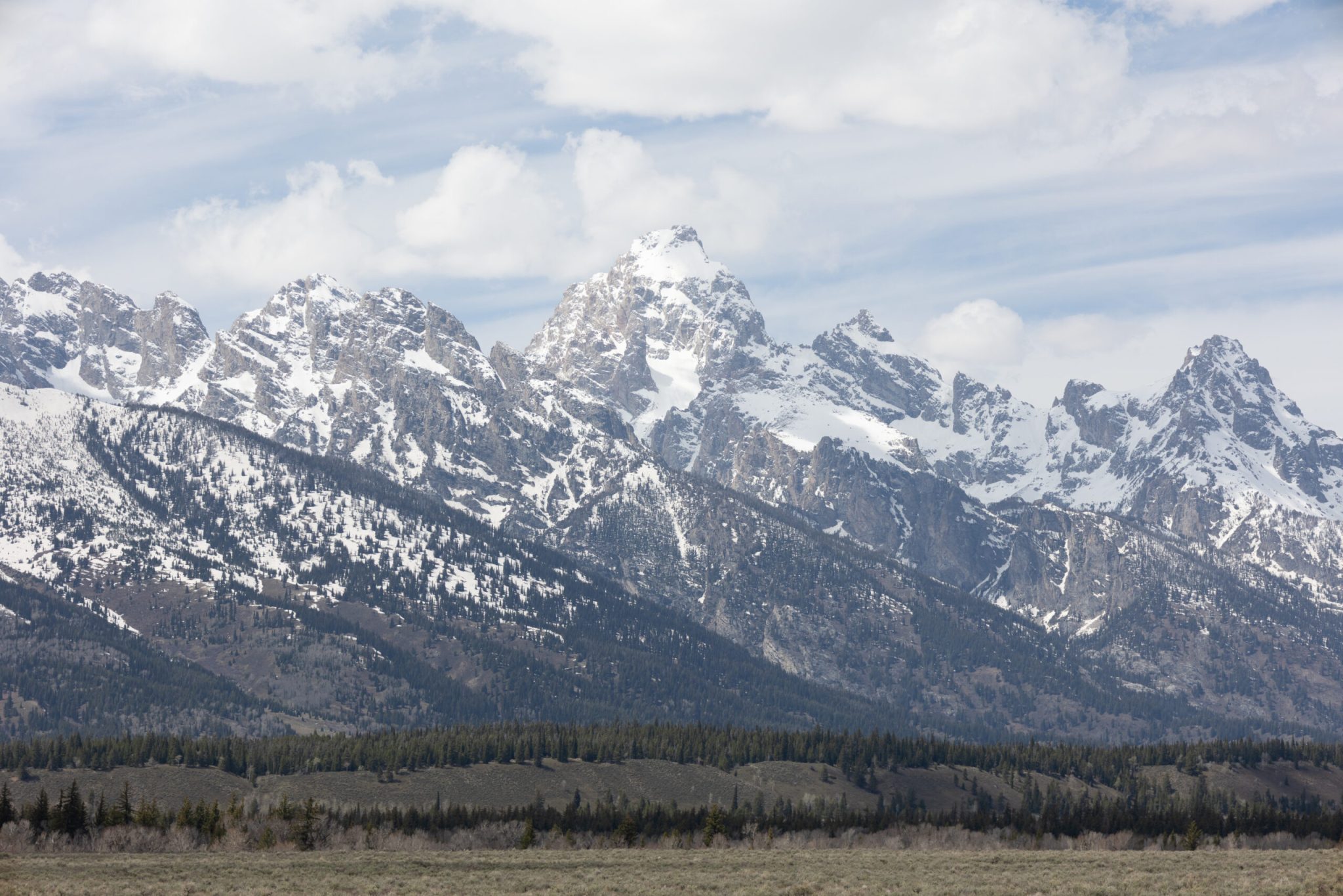 Scenic view of the snow-capped Grand Tetons, photographed by Preston Ackerman, featuring a lush green forest at the base of the rugged mountains under a cloudy sky.