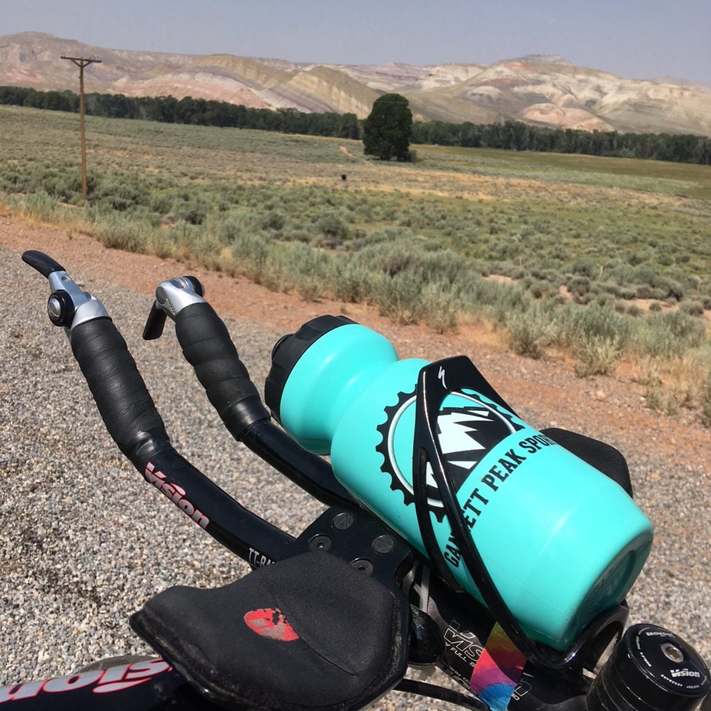 A turquoise water bottle with a mountain logo is attached to the handlebars of a bicycle, with a vast desert landscape stretching out behind it in Wind River Country.