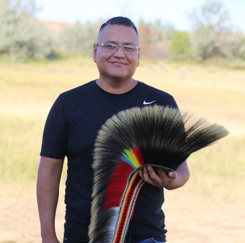 A man stands in a grassy field holding a colorful feathered headdress in Wind River Country.