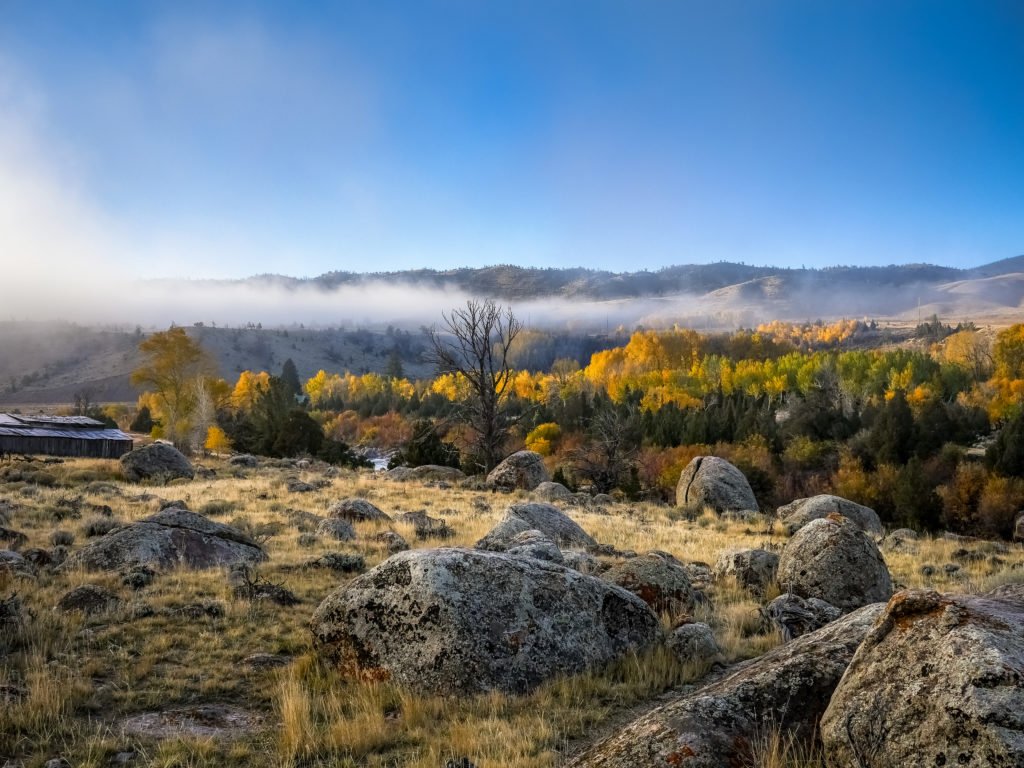 A vast, open landscape with scattered rocks and patches of dry grass, leading up to a distant mountain range shrouded in mist, under a clear blue sky in Wind River Country.