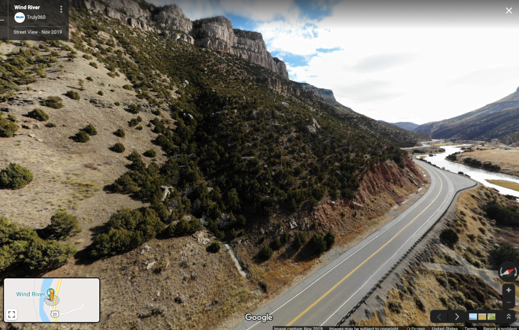 A winding highway cuts through a rugged, mountainous landscape with sparse vegetation and a river flowing nearby in Wind River Country.