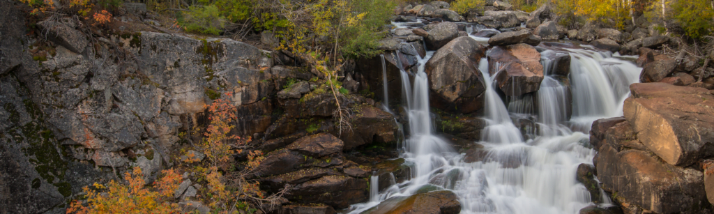A cascading waterfall flows over rugged, moss-covered rocks, surrounded by autumn foliage and evergreen trees in Wind River Country.