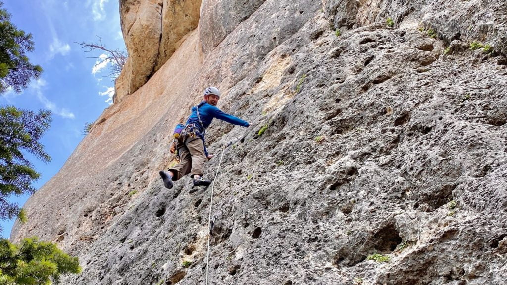 A rock climber ascends a steep, rugged cliff face with a clear blue sky above and trees in the background in Wind River Country.