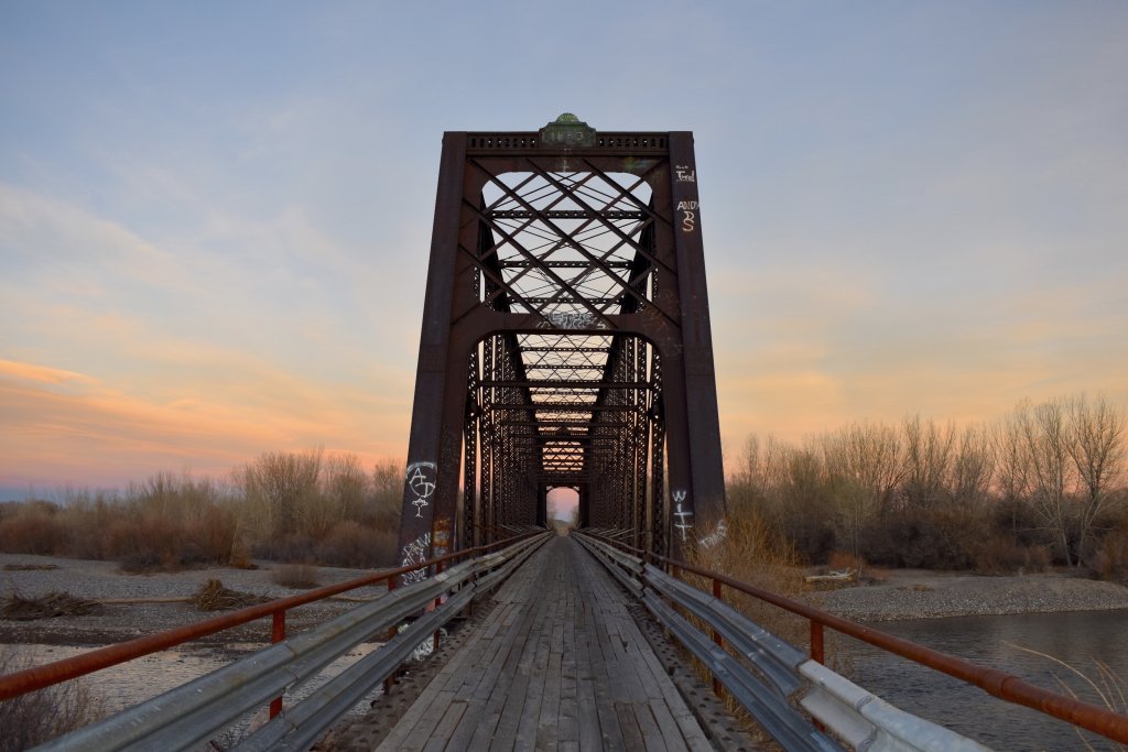 A rusted metal truss bridge with a wooden deck spans a river at sunset, surrounded by bare trees and a serene landscape in Wind River Country.