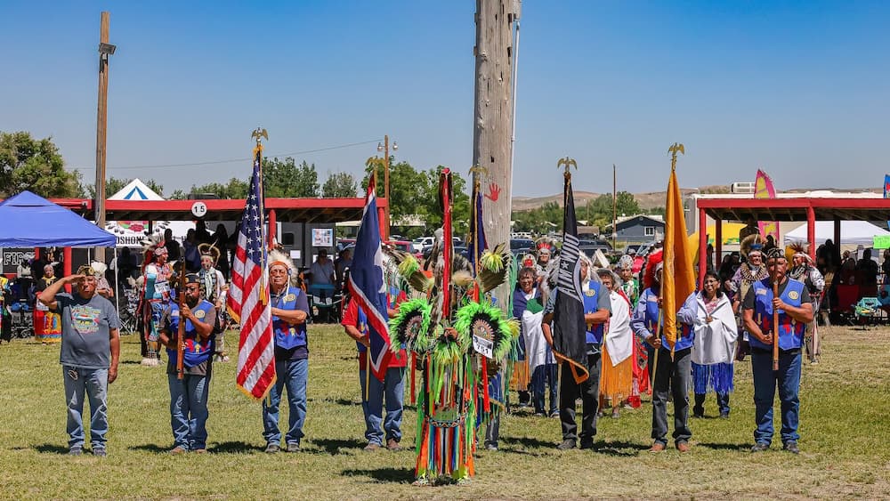 Nine Things to Do on the Wind River Indian Reservation A vibrant gathering of Native American Powwow community members in traditional regalia at a cultural event in Wind River, Wyoming, with flags held proudly under a clear blue sky.