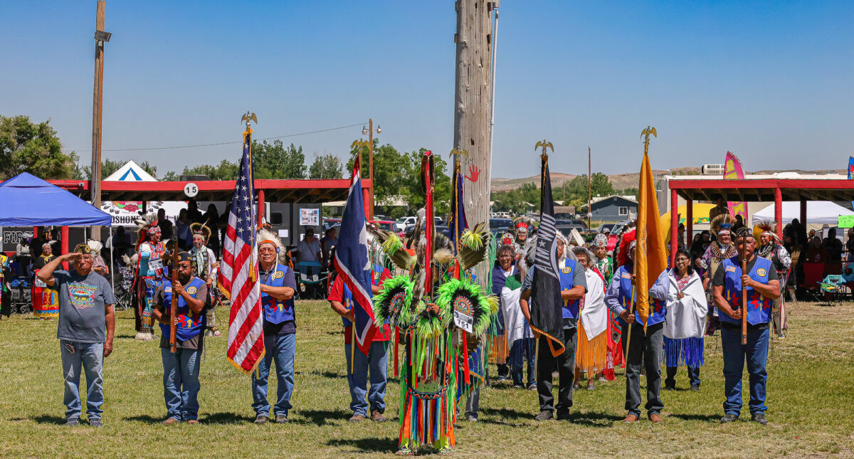 A vibrant gathering of Native American Powwow community members in traditional regalia at a cultural event in Wind River, Wyoming, with flags held proudly under a clear blue sky.
