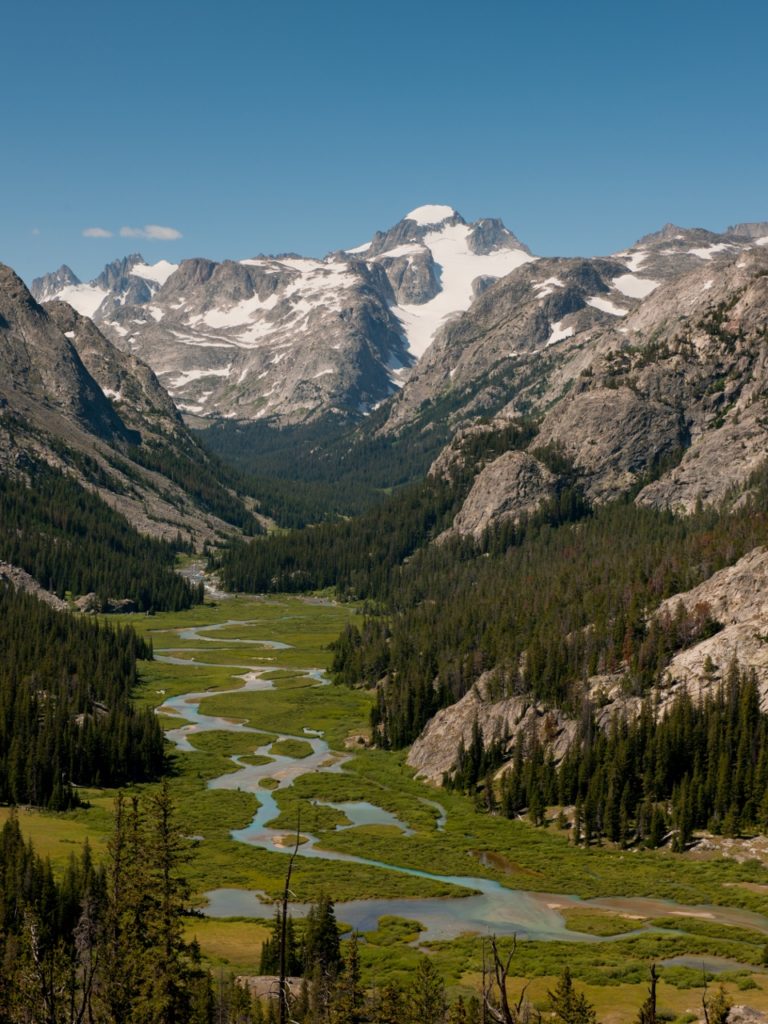 A serene mountain valley with a winding river, lush green meadows, and towering snow-capped peaks under a clear blue sky in Wind River Country.