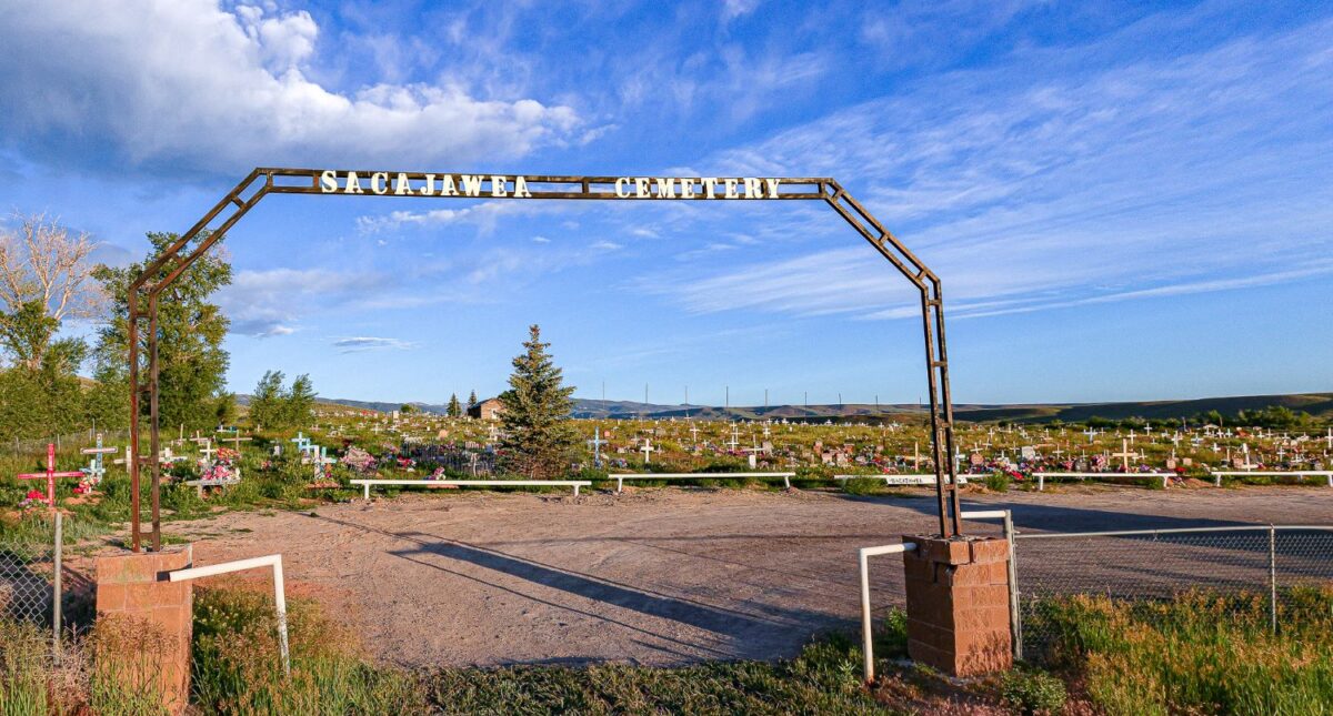 The entrance to Sacajawea Cemetery in Wind River, Wyoming, with a metal archway sign reading 'Sacajawea Cemetery' against a backdrop of decorated graves.