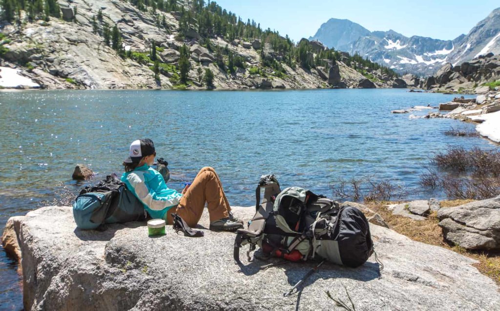 A hiker takes a break on a rocky shore, overlooking a serene alpine lake with snow-capped mountains in the distance in Wind River Country.