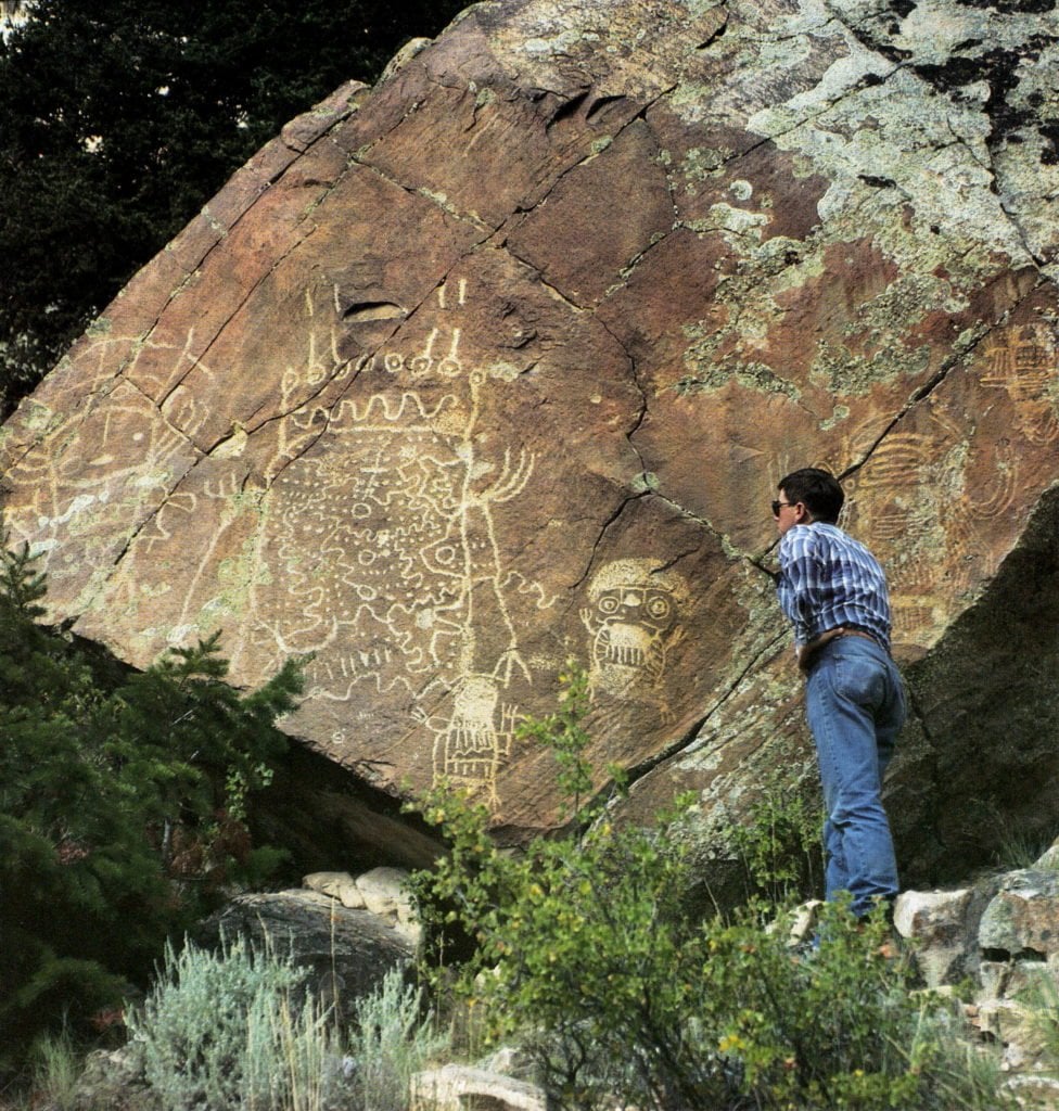 A person examines ancient petroglyphs on a massive rock face in a forested area in Wind River Country.