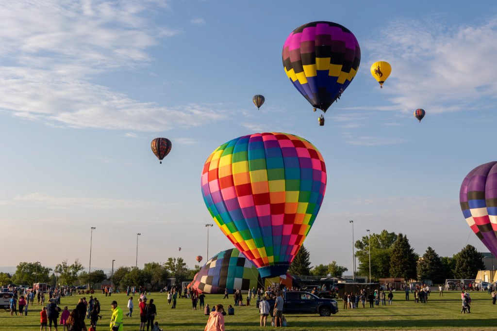 A vibrant hot air balloon festival with colorful balloons ascending into a clear blue sky over a grassy field filled with spectators in Wind River Country.