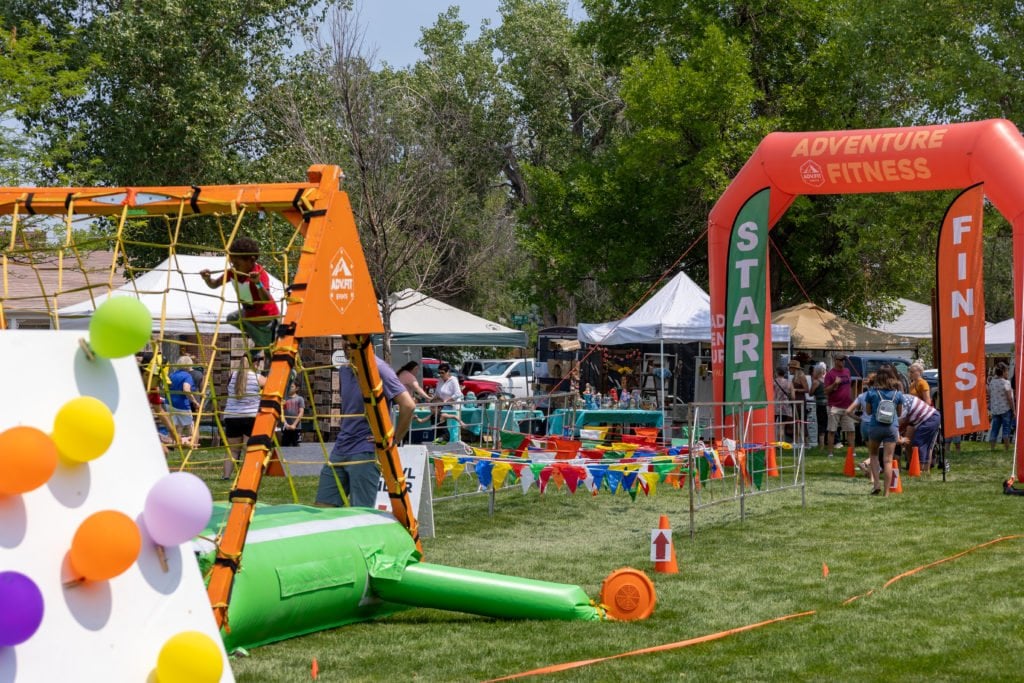 A vibrant outdoor event with colorful inflatable structures, balloons, and tents set up in a grassy area, featuring an Adventure Fitness race with participants and spectators in Wind River Country.