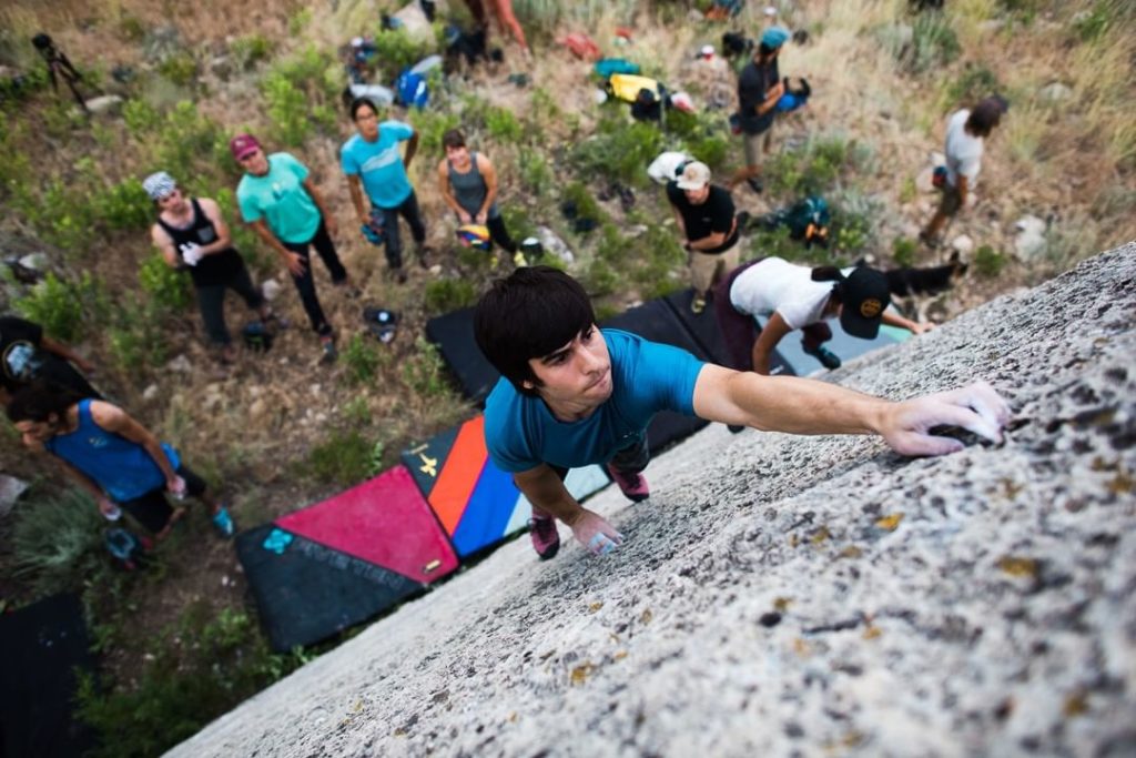 A young man in a blue shirt and climbing shoes ascends a steep rock face, reaching for a hold, while a group of spectators watches from below in Wind River Country.