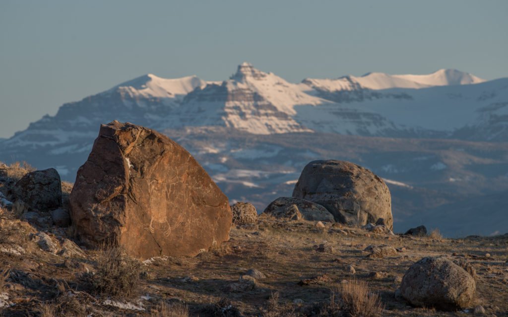 A rugged landscape with large boulders and sparse vegetation in the foreground, leading up to majestic snow-capped mountains in the distance in Wind River Country.