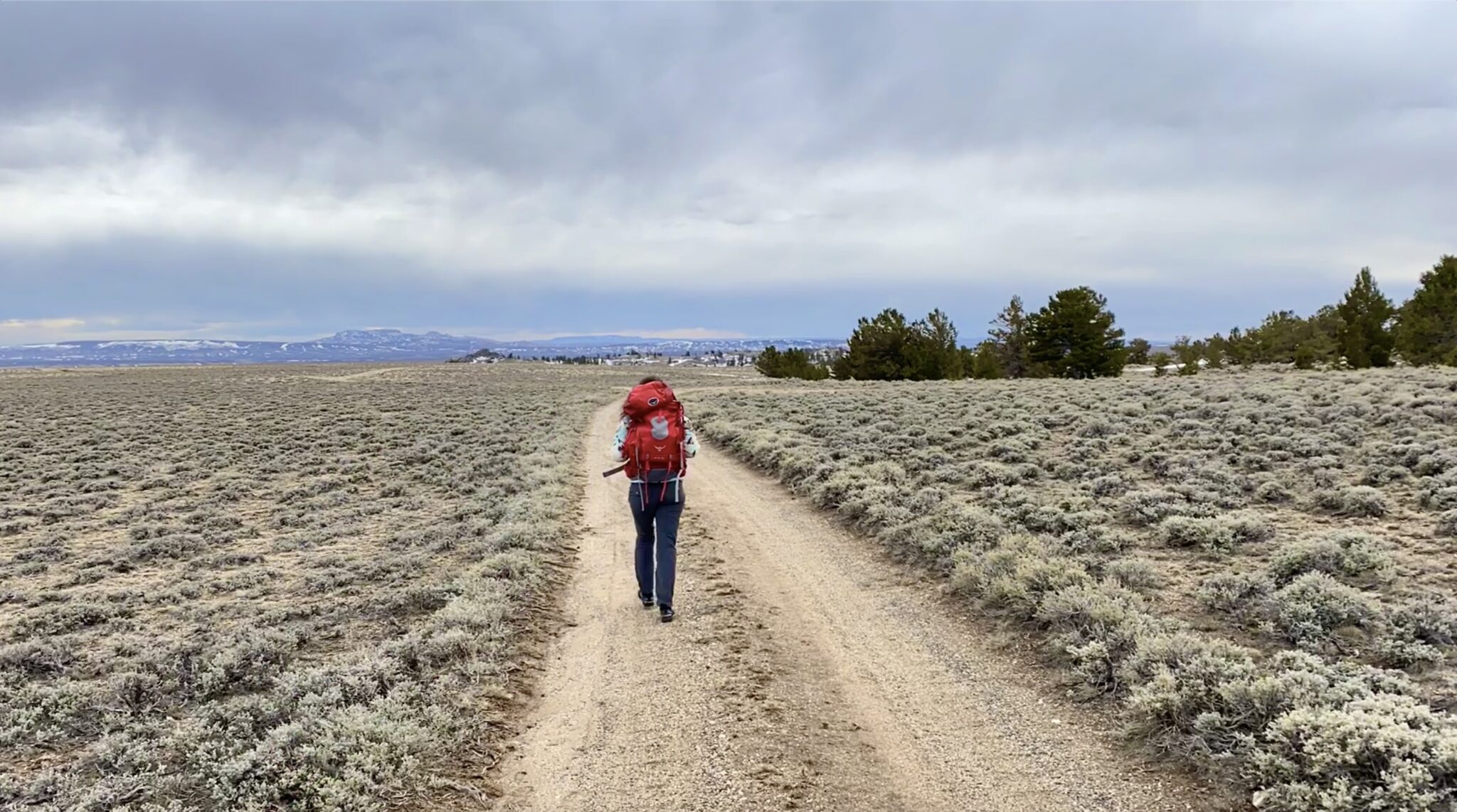 A hiker with a red backpack walks along a winding dirt path through a vast, dry landscape dotted with sparse vegetation in Wind River Country.