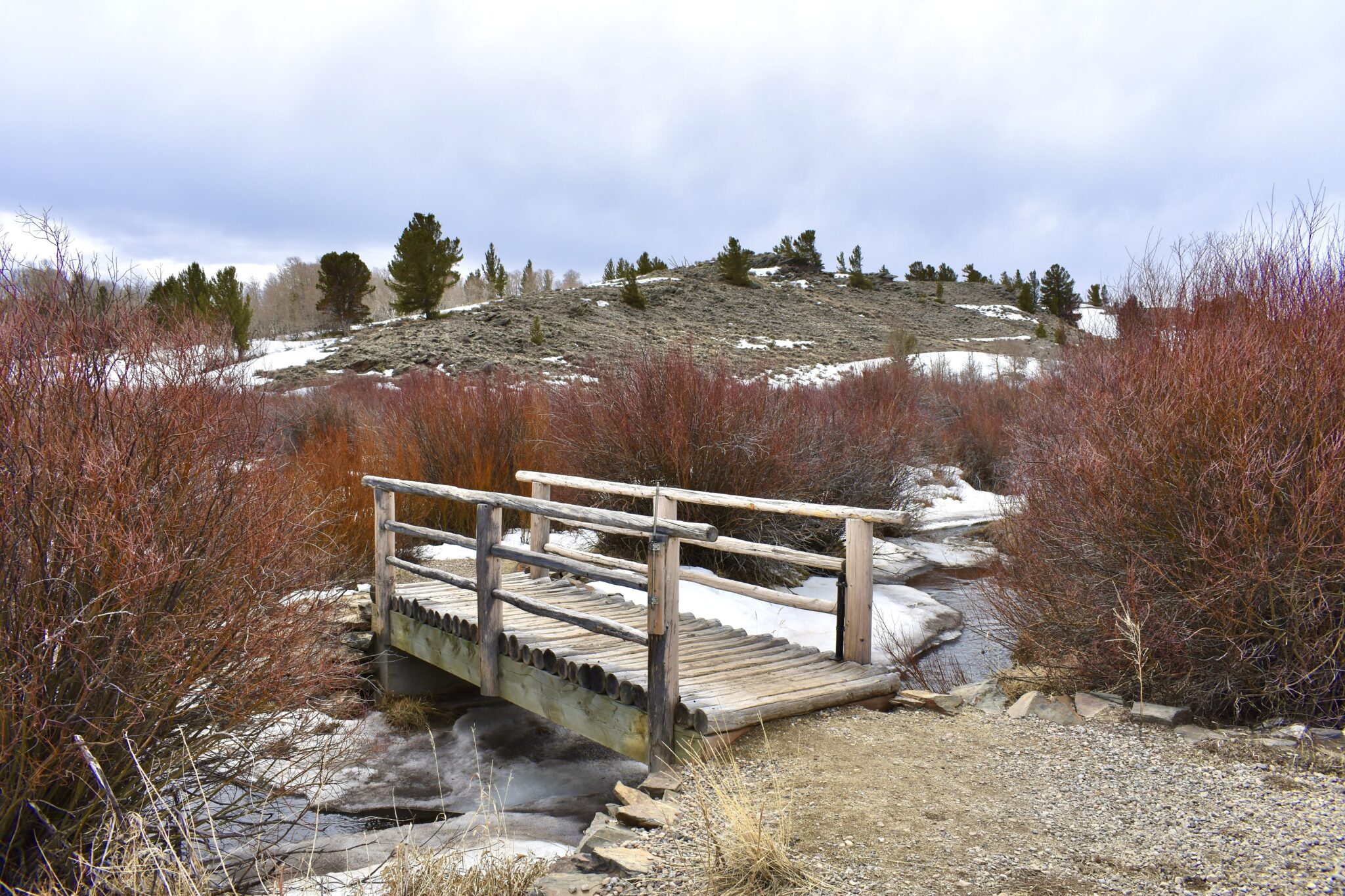 A rustic wooden bridge spans a small stream in a wintry landscape, surrounded by snow-dusted hills and bare trees in Wind River Country.