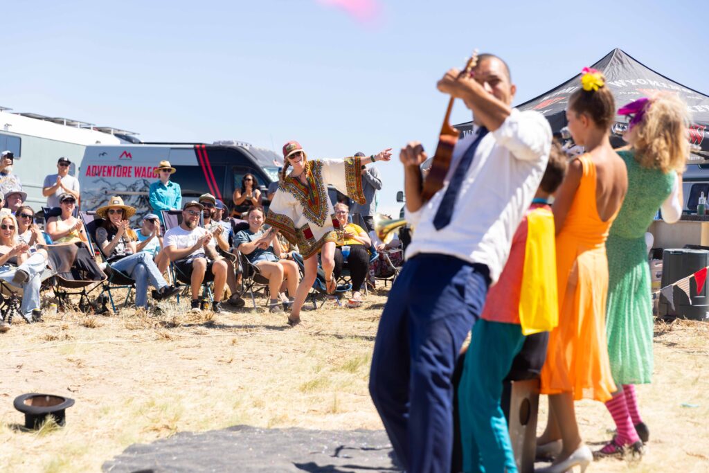 A lively outdoor performance with a musician playing guitar, accompanied by colorful dancers, while an audience enjoys the show under a clear blue sky in Wind River Country.