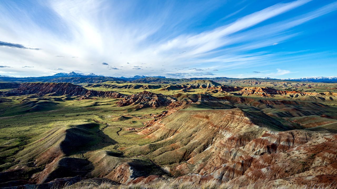 A vast desert landscape with colorful rock formations and distant snow-capped mountains under a clear blue sky in Wind River Country.