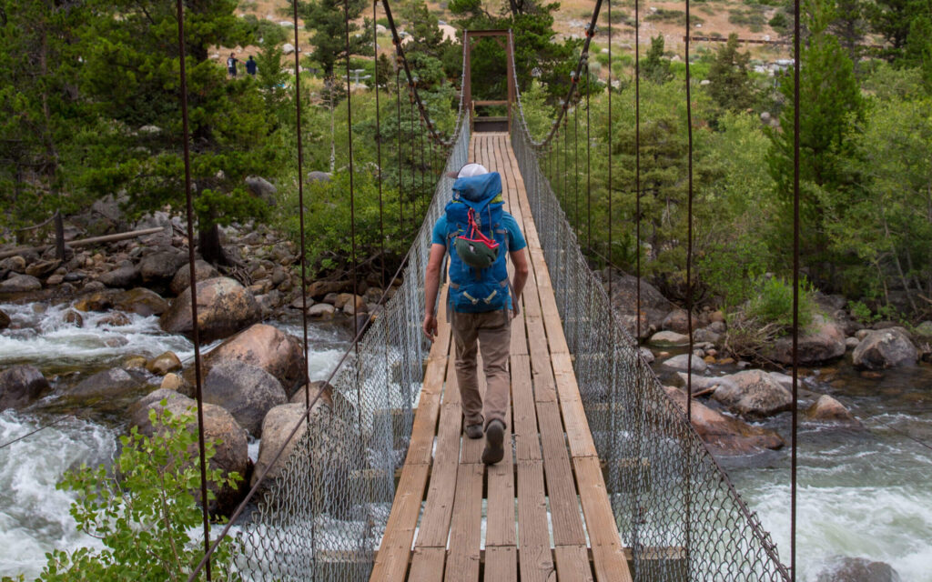 Hiker traversing a suspension bridge over a rushing river in Sinks Canyon, Wind River Range, emphasizing the thrill of hiking adventures.