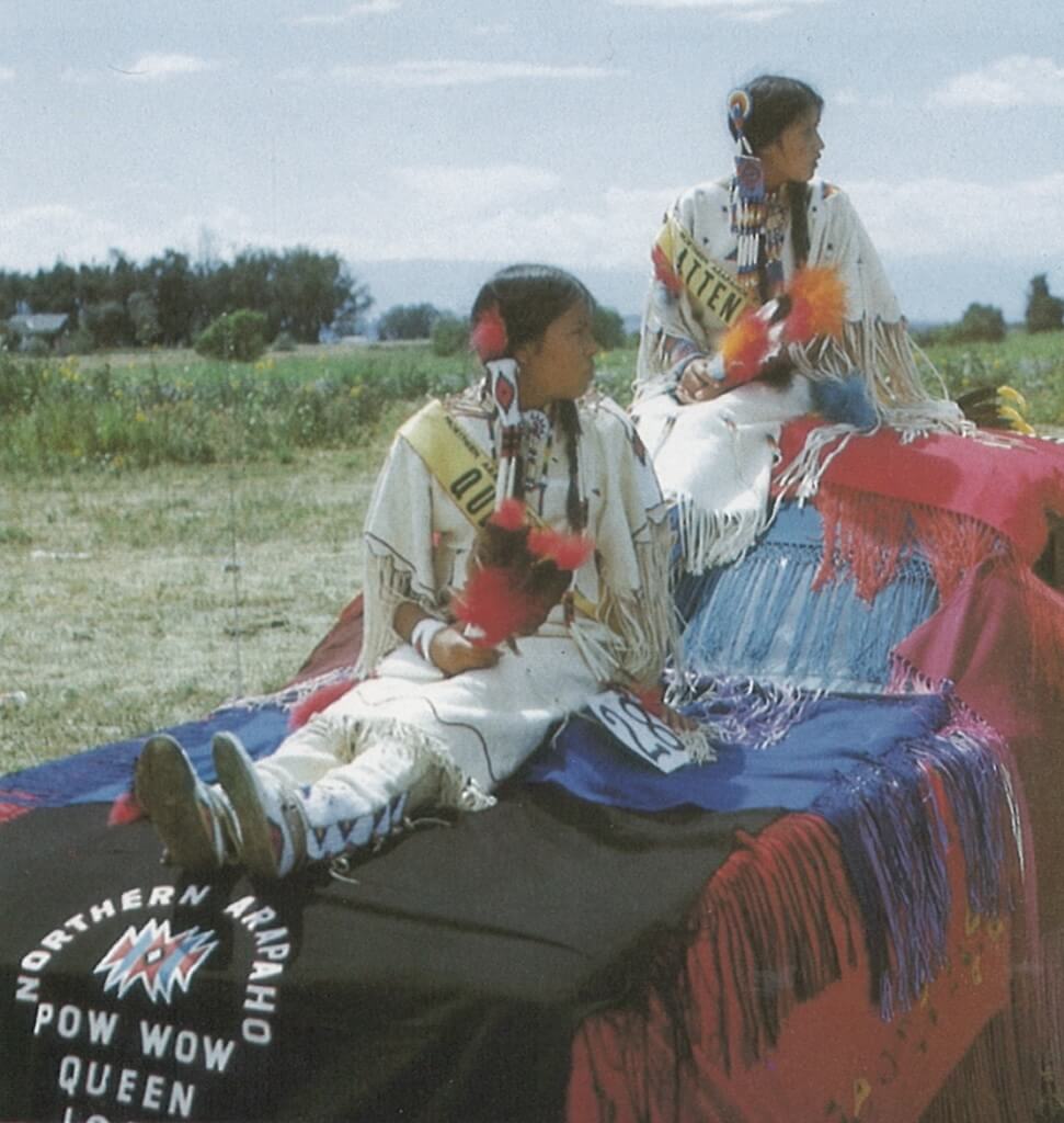 Two young Native American women in traditional regalia sitting on a parade float in Wind River Country.