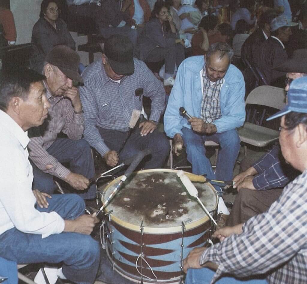 A group of men sitting in a circle playing a large drum together in Wind River Country-Arapaho Eagle Drum Group.