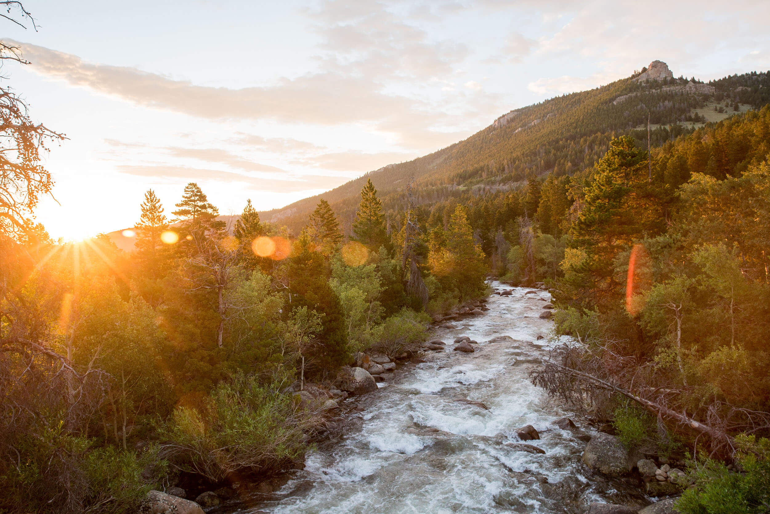 Sunset over a river in Wyoming with lush greenery and mountains in the background. Credit: Wyoming Office of Tourism.