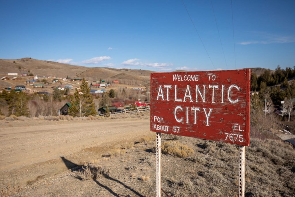 A rustic wooden sign welcomes visitors to Atlantic City, a small town nestled in a valley surrounded by rolling hills and scattered houses in Wind River Country.