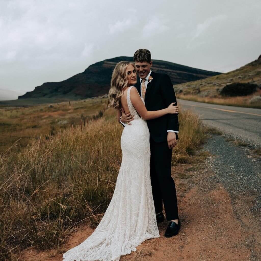 A bride and groom embrace on a roadside with mountains in the background in Wind River Country.