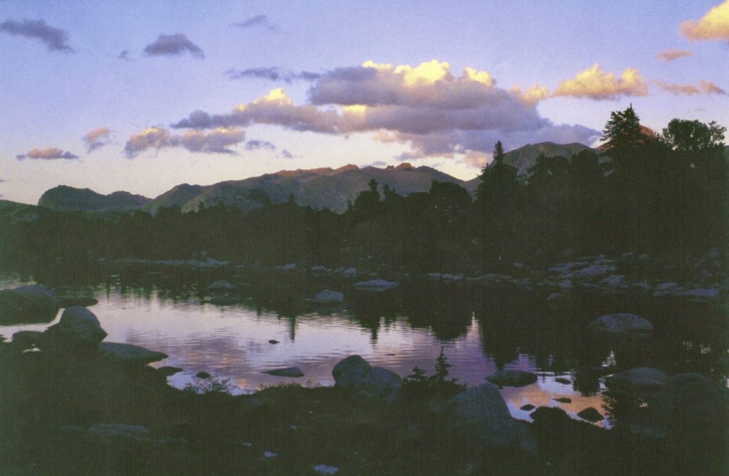 A serene mountain lake at dusk, surrounded by rocky shores and silhouetted trees, with a reflection of the colorful sky on the water's surface in Wind River Country.