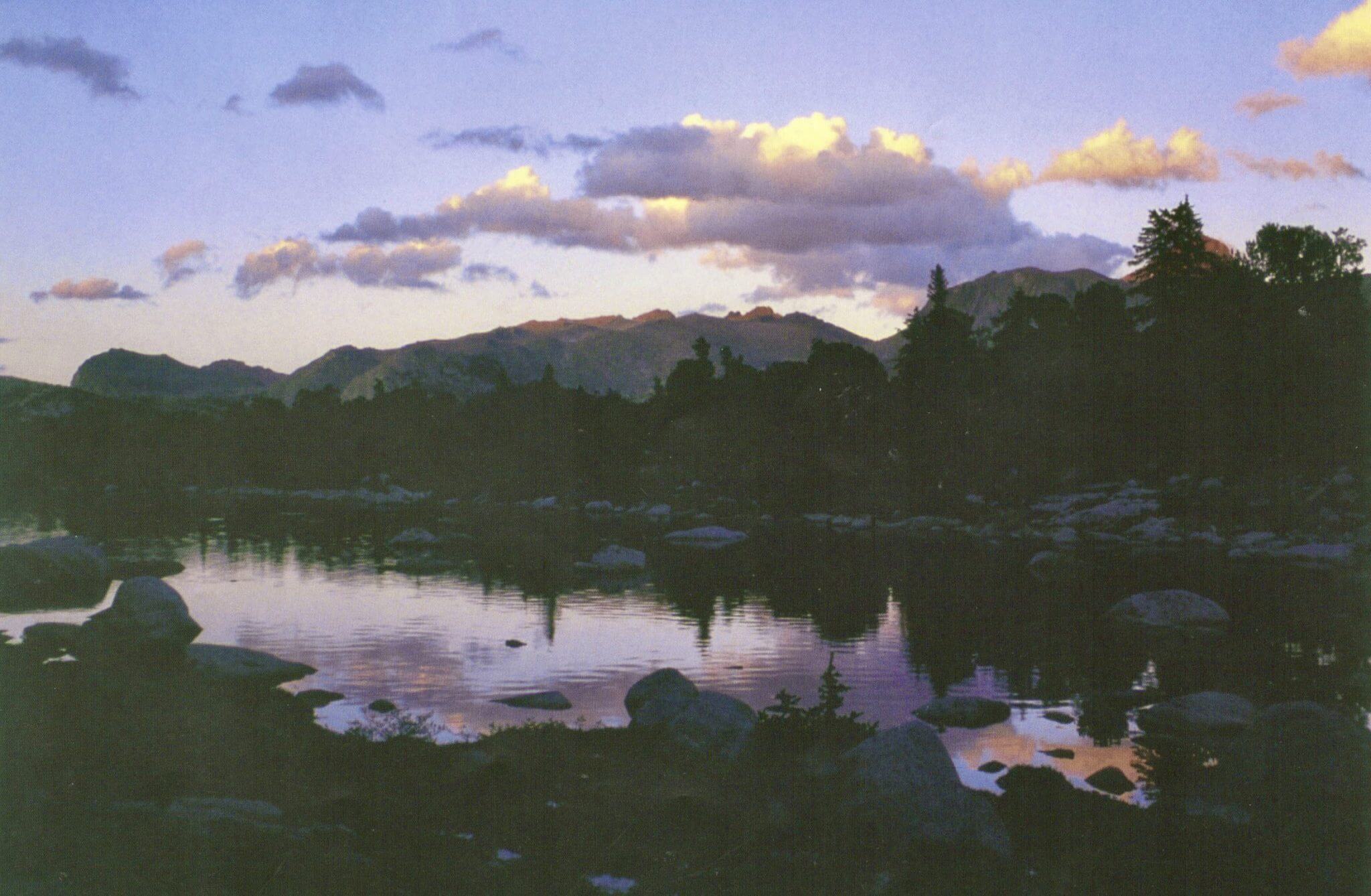 A serene mountain lake at dusk, surrounded by rocky shores and silhouetted trees, with a reflection of the colorful sky on the water's surface in Wind River Country.