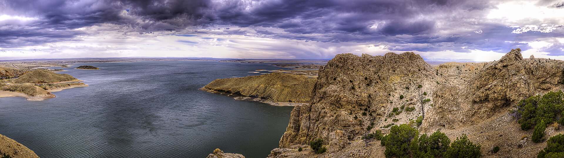 A panoramic view of a vast lake surrounded by rugged, rocky cliffs and sparse vegetation under a dramatic, stormy sky in Wind River Country.